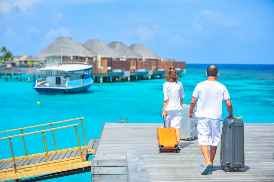 Travelers walking with luggage toward overwater bungalows at a tropical resort